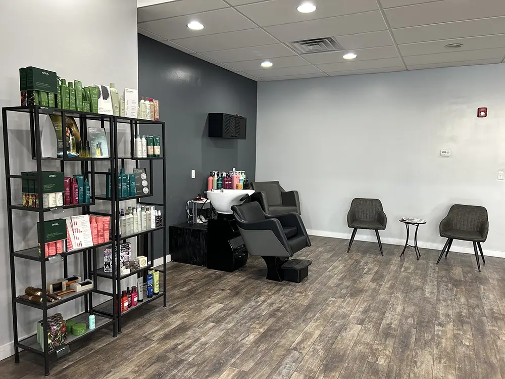 Modern salon interior with two hair wash stations, a product display shelf on the left, and a small waiting area with two chairs and a table on the right. - Styles by Stacy - Poughkeepsie, NY