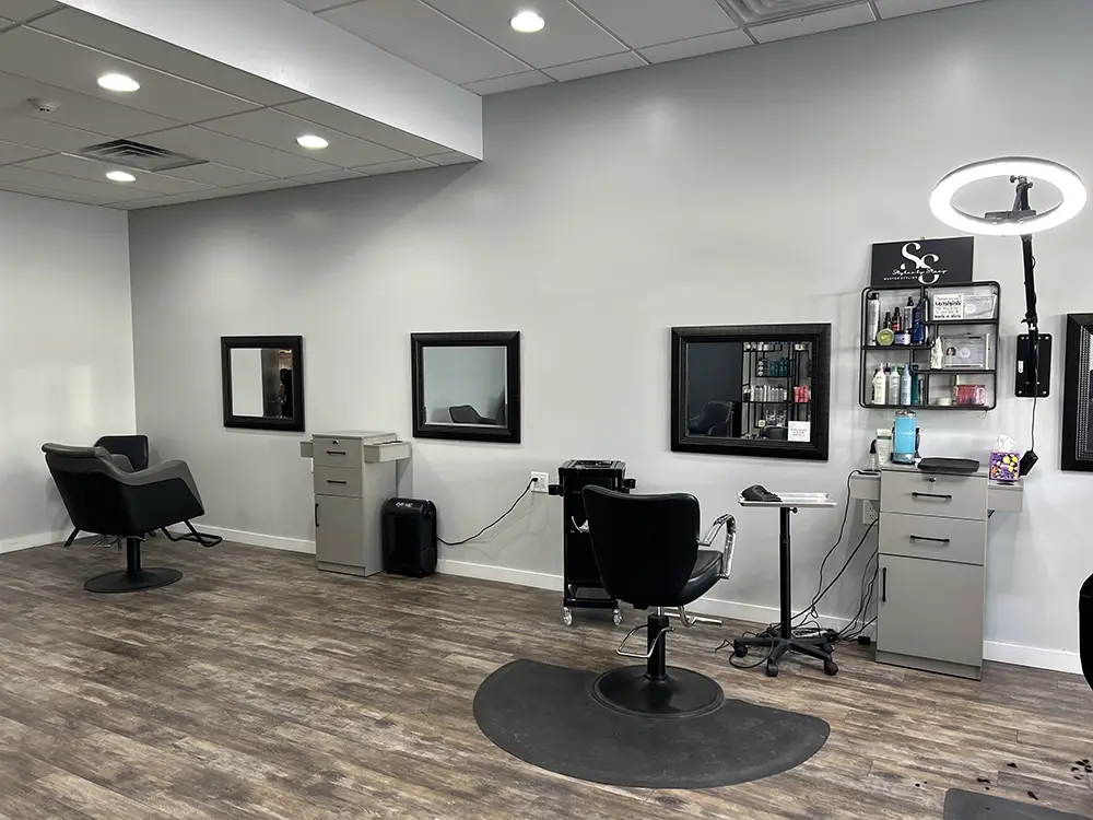 A modern hair salon with three black styling chairs, mirrors, gray storage cabinets, shelves with hair products, and wood-look flooring. - Styles by Stacy - Poughkeepsie, NY