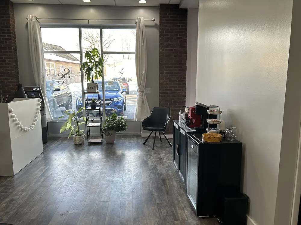 A modern waiting area with large windows, a black chair, potted plants, a small refreshment station, and parked cars visible outside. - Styles by Stacy - Poughkeepsie, NY